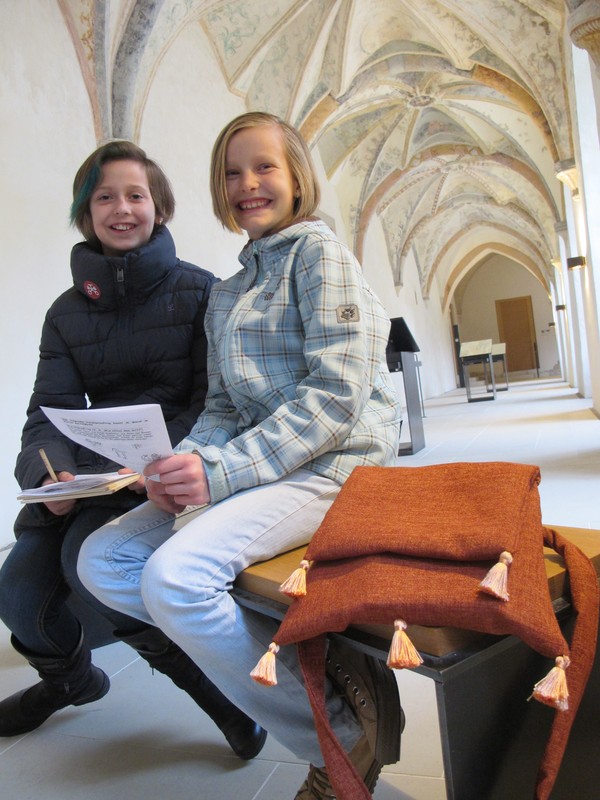 Zwei Kinder sitzen mit einer Pilgertasche auf einer Bank im Kreuzgang und lachen.