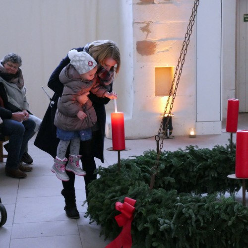 Ein kleines Mädchen und eine Erwachsene entzünden eine Kerze auf einem sehr großen Adventskranz. Im Hintergrund sitzen Menschen.