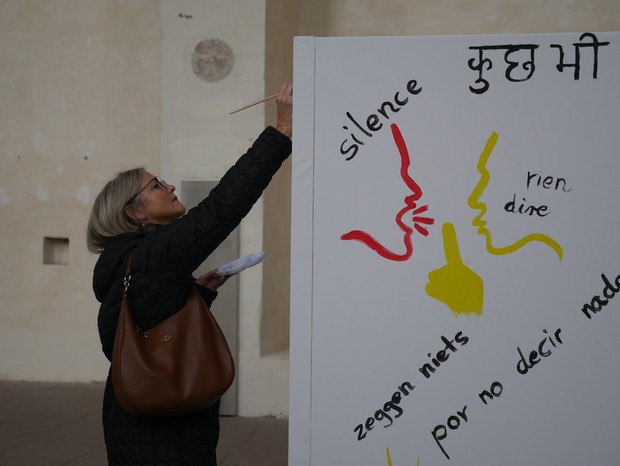 Eine Frau malt auf eine Leinwand. Darauf sind Schweigesymbole und unterschiedliche Sprachen gemalt.