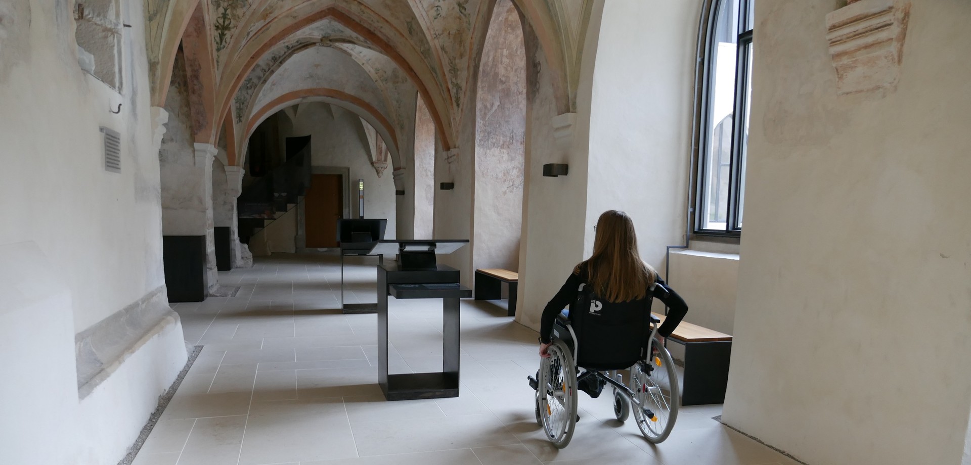 Eine Frau fährt im Rollstuhl durch den Kreuzgang und sieht sich um.