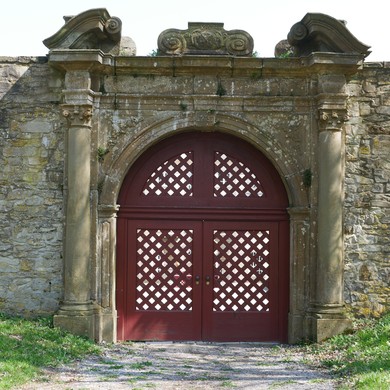 Ein rotes, zweiflügeliges Tor befindet sich in einer alten Mauer. Der Torbogen aus Sandstein ist reich verziert.