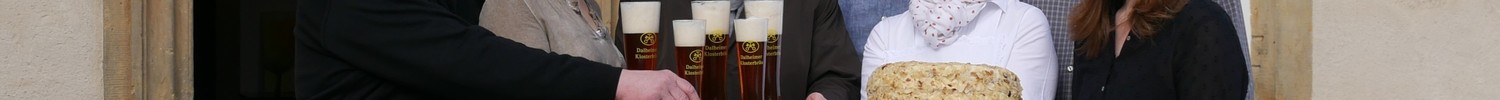 Eine Gruppe von Menschen steht vor dem Wirtshaus und hält einen Kuchen und Biergläser.