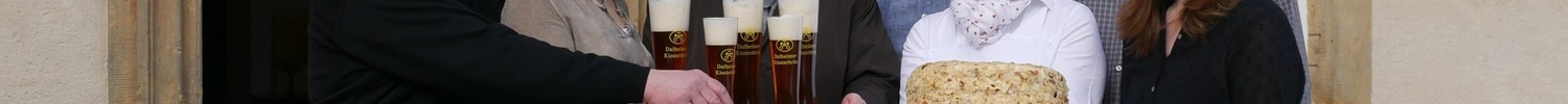 Eine Gruppe von Menschen steht vor dem Wirtshaus und hält einen Kuchen und Biergläser.