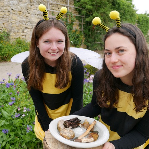 Zwei als Bienen verkleidete junge Frauen stehen an einer Insektentränke und schauen freundlich in die Kamera.