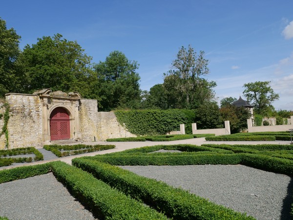 Ein Garten mit symmetrischen Kiesflächen und kleinen Hecken. Dahinter steht eine alte Mauer, in die ein großes, rotes Tor eingelassen wurde.