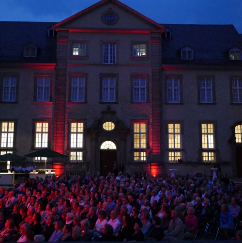 Das Publikum der Sommernachtslieder sitzt im Ehrenhof. Es ist bereits dunkel, die Fassade des alten Gebäudes und stimmungsvoll beleuchtet.