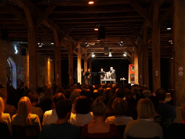 Im abgedunkelten Schafstall sitzt das Publikum vor einer beleuchtetend Bühne mit Musikern und einer lesenden Person. Im abgedunkelten Schafstall sitzt das Publikum vor einer beleuchtetend Bühne mit Musikern und einer lesenden Person.