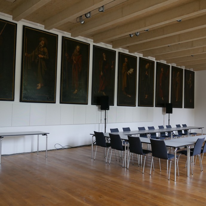 Blick in den Vortragsraum. In der Mitte stehen Tische in Hufeisenform, an der Wand hängen große Gemälde. (vergrößerte Bildansicht wird geöffnet)