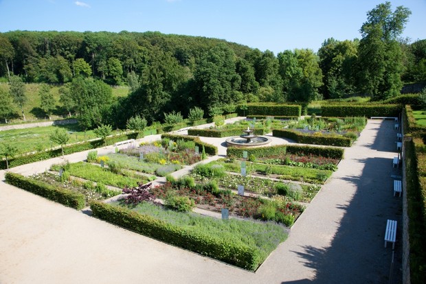 Ein Garten mit mehreren kleinen, symmetrisch angelegte Beeten und Hecken. In der Mitte befindet sich ein Springbrunnen.