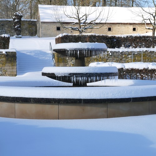 In einem verschneiten Garten steht ein Springbrunnen, an sich viele Eiszapfen gebildet haben. Im Hintergrund befinden sich Hecken, Bäume und ein kleines Gebäude.