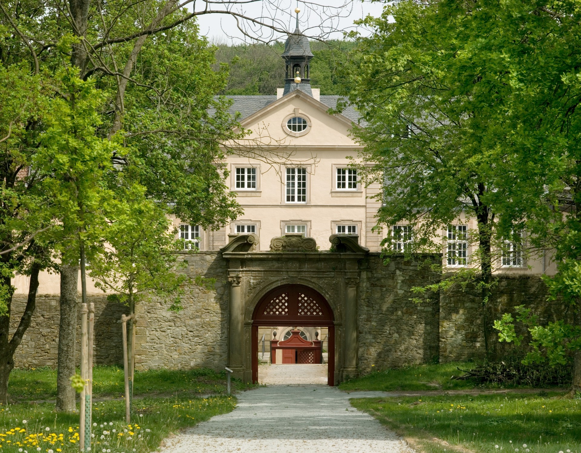 Ein Weg führt auf eine geöffnete, reich verzierte Tordurchfahrt zu. Dahinter befindet sich ein historisches Gebäude.