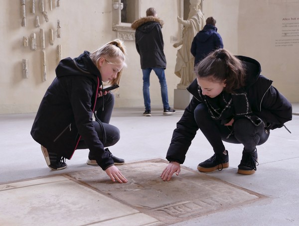 Zwei Mädchen tasten den Boden einer Kirche ab, in den etwas eingraviert ist. Im Hintergrund sind weitere Personen.