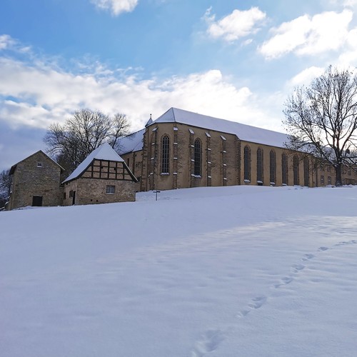 Hinter einer schneebedeckten Anhöhe befindet sich die gotische Klosterkirche Dalheims. Links davor stehen zwei kleine Häuser aus hellem Bruchstein.