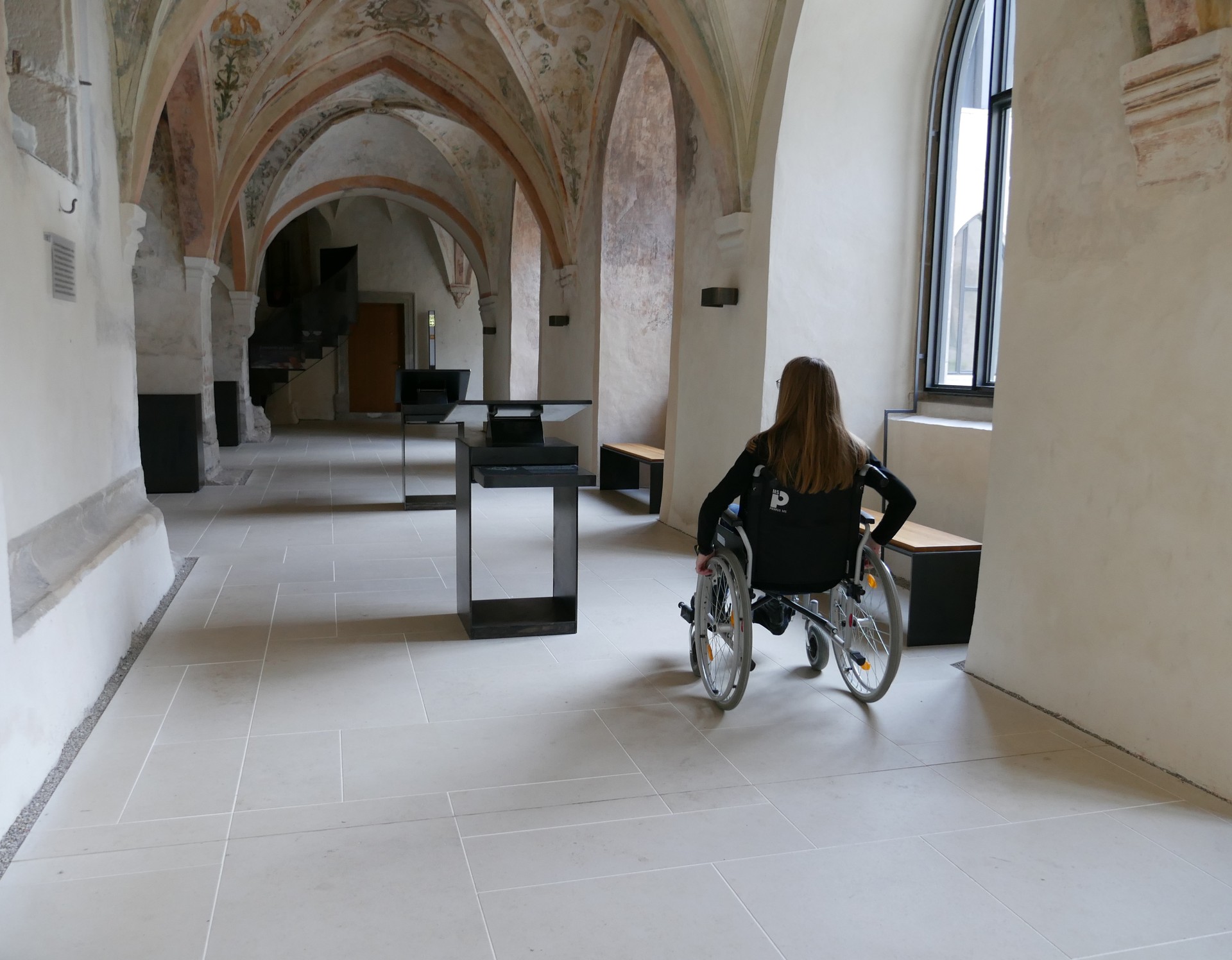 Eine Frau fährt im Rollstuhl durch den Kreuzgang und sieht sich um.