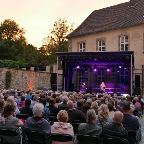 Ein Mann steht im Ehrenhof bei Sonnenuntergang vor Publikum auf einer Bühne und singt.