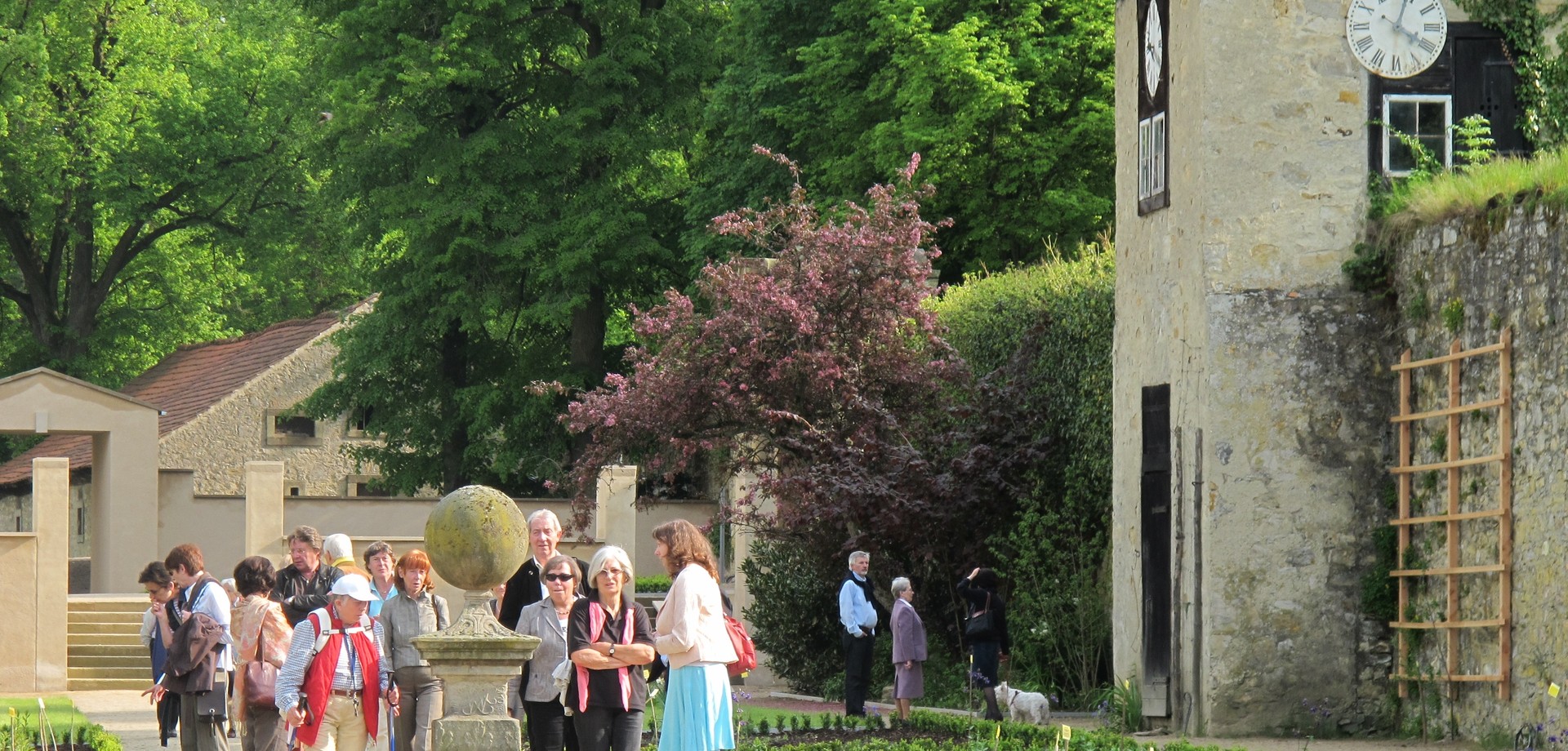In einem Garten staht eine Gruppe um eine Steinskulptur. Rechts befindet sich ein kleiner Uhrturm.