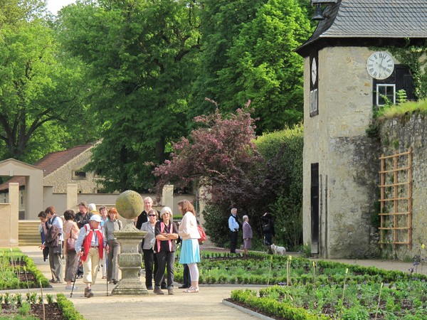 In einem Garten staht eine Gruppe um eine Steinskulptur. Rechts befindet sich ein kleiner Uhrturm.