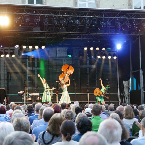 Drei Frauen in grünen Kleidern stehen vor Publikum auf einer Bühne und halten ihre Instrumente in die Höhe.