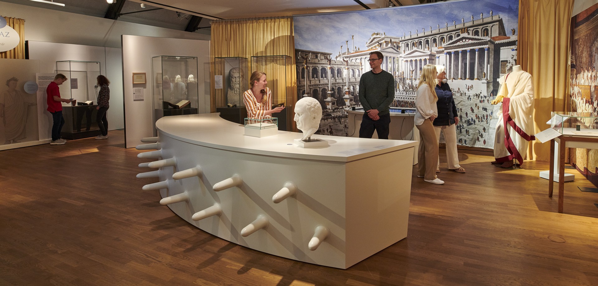 Mehrere Menschen schauen sich im Ausstellungsraum von "Latein. Tot oder lebendig!?" die Exponate an.