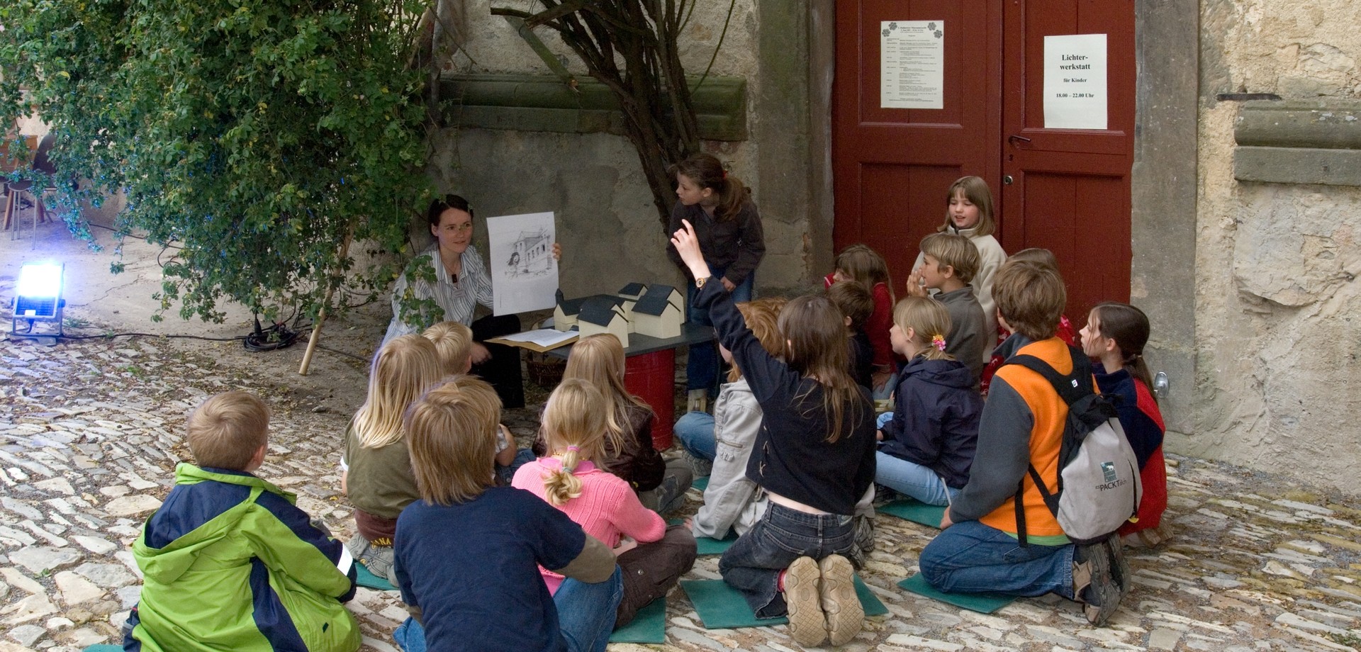 Eine Gruppe von Kindern sitzt in einem Hof. Eine Frau hält ein Bild hoch. Vor ihr stehen einige kleine Modellhäuser. Ein Mädchen hebt den Arm.