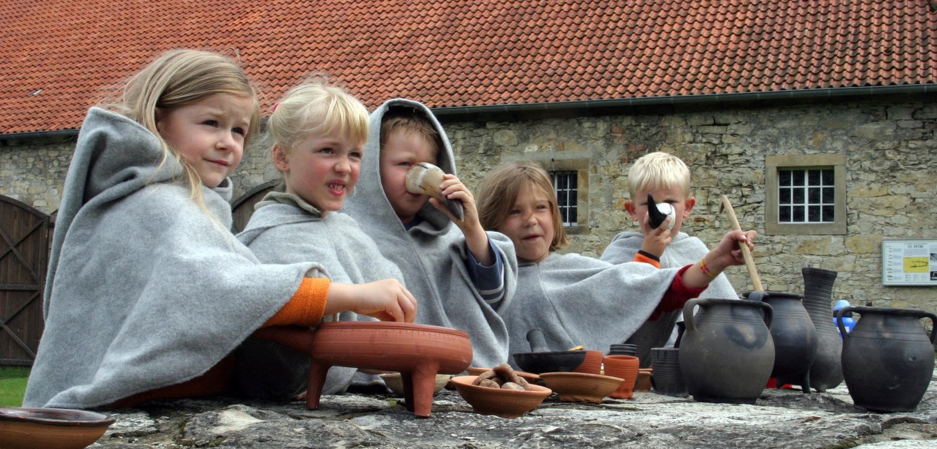 Kinder in grauen Umhängen sitzen an einem Steinblock. Vor ihnen stehen verschiedene Tongefäße, in denen sie rühren. Zwei der Kinder trinken aus Trinkhörnern.